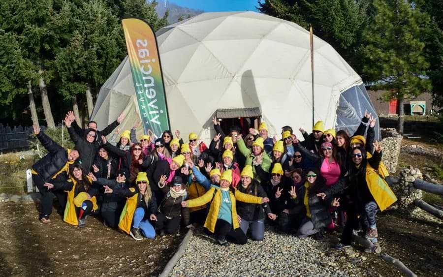 Grupo grande de jovens comemorando diante de um domo, com bandeiras da Vivencias Travel