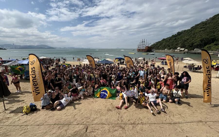Grupo numeroso de estudantes na praia, junto às bandeiras da Vivencias Travel