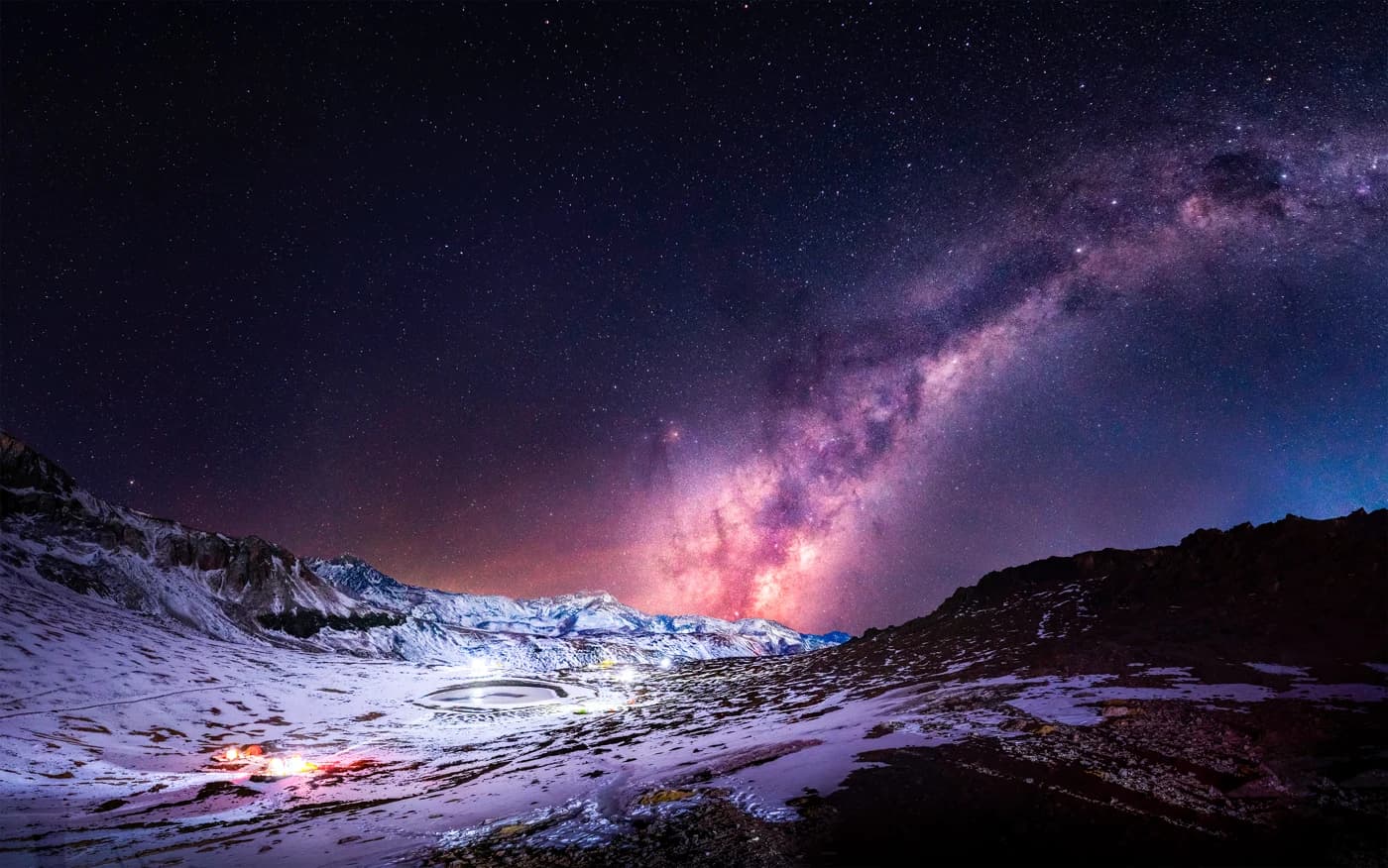 O centro da Via Láctea sobre a cordilheira nevada do Chile, com uma laguna congelada e luzes de acampamento no vale
